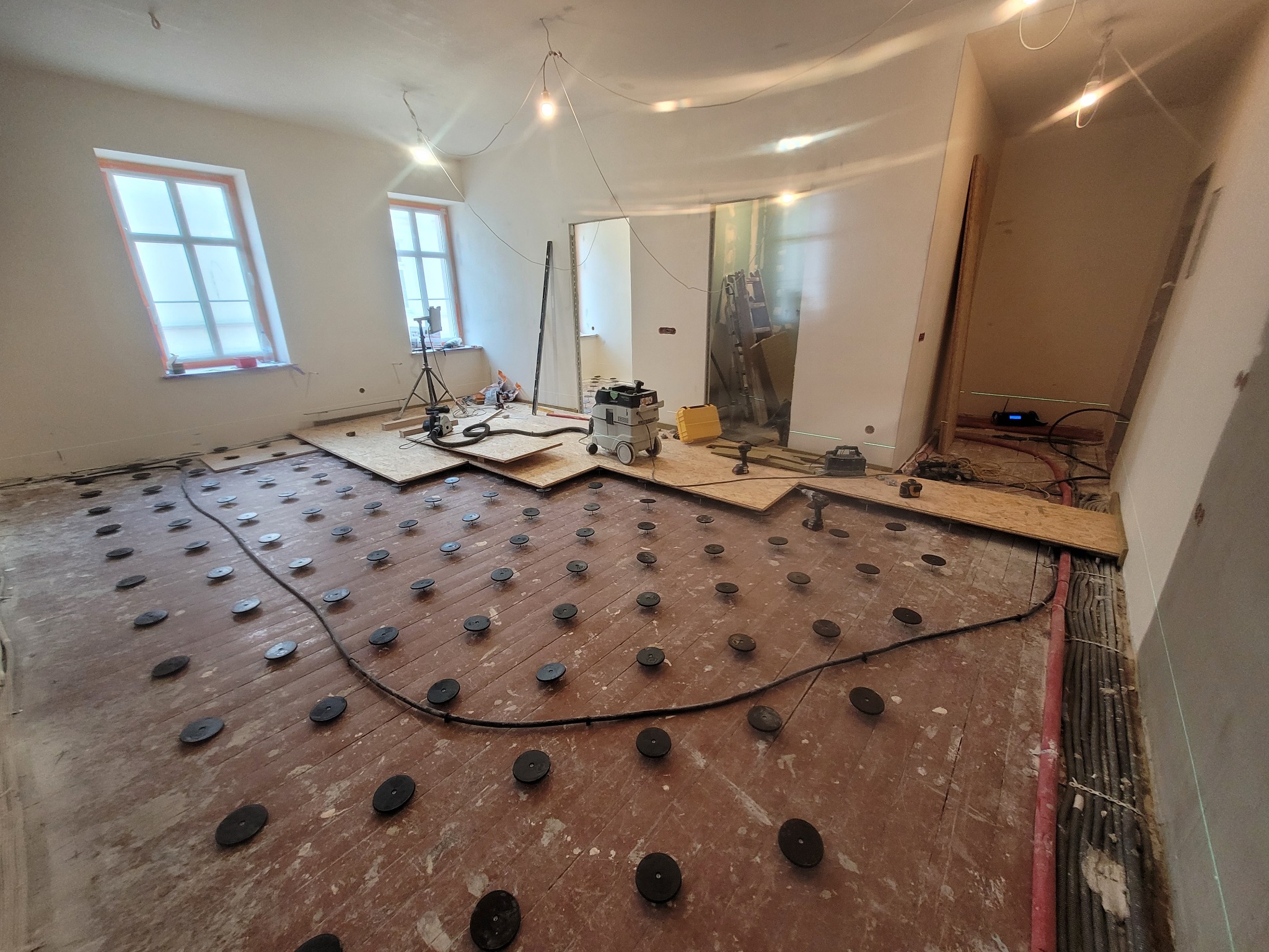 Przygotowanie podłoża pod podłogę w kamienicy: widoczne płyty OSB, system poziomowania Atlas M-system i narzędzia. W tle okna i oświetlenie budowlane.