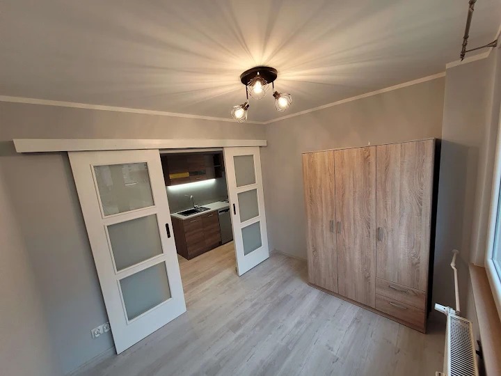 Jasne wnętrze po remoncie: przesuwne drzwi z matowego szkła, drewniana szafa, szare ściany, nowoczesna lampa sufitowa. Widok na aneks kuchenny.