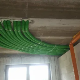 Zielone rury wentylacyjne zamontowane pod betonowym sufitem, zabezpieczone pianką montażową, widoczne mocowania i fragment pomarańczowej rury.