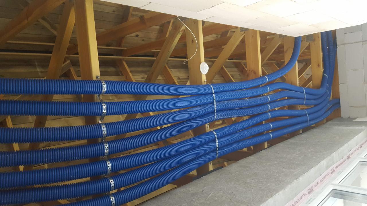 Niebieskie karbowane przewody wentylacyjne zamocowane do drewnianej konstrukcji stropu za pomocą metalowych opasek zaciskowych, widoczne bloczki betonowe i fragment okna.