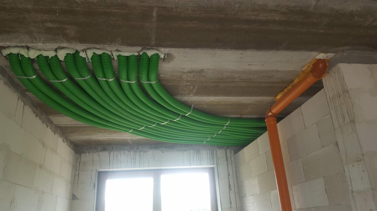 Zielone rury wentylacyjne zamontowane pod betonowym sufitem, zabezpieczone pianką montażową, widoczne mocowania i fragment pomarańczowej rury.