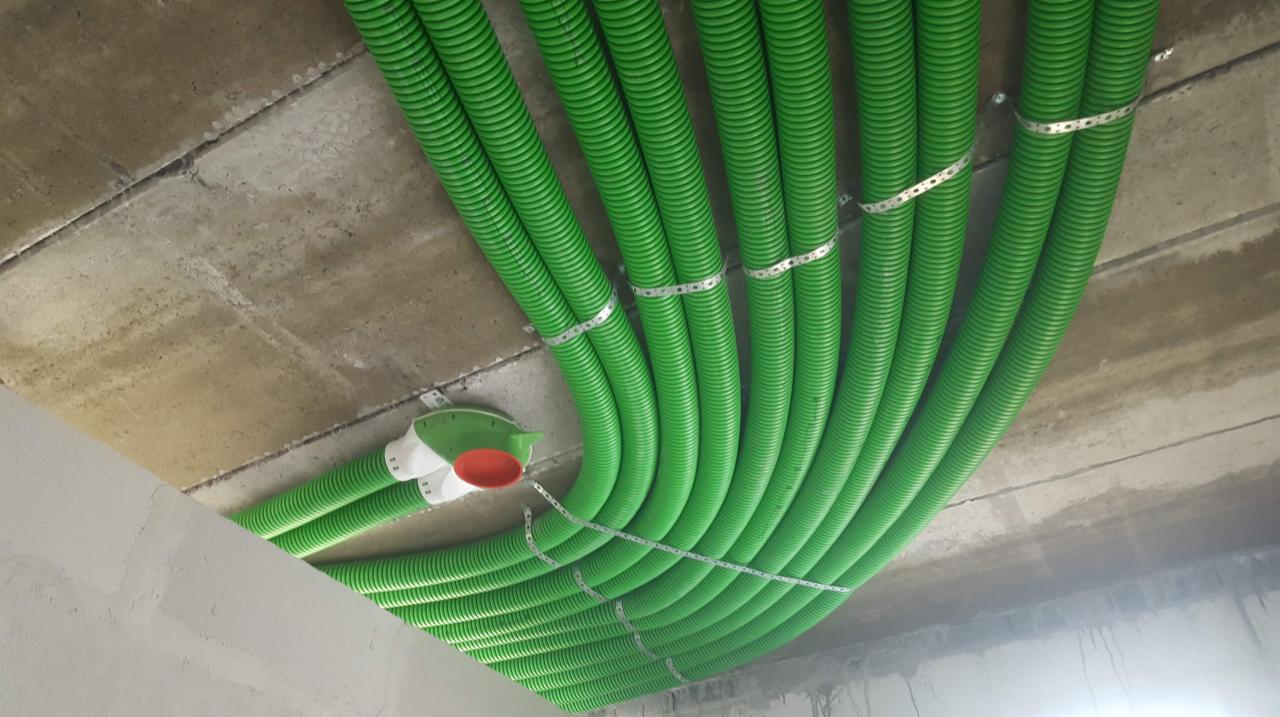 Zielone, karbowane rury przymocowane metalowymi obejmami do sufitu z betonu architektonicznego, widoczna puszka rozdzielcza z pomarańczową pokrywą.