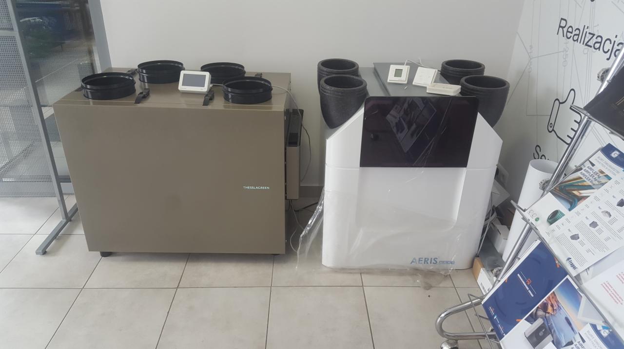 Dwa urządzenia do wymiany powietrza: szare 'THESSLAGREEN' i białe 'AERIS', z czarnymi okrągłymi wlotami powietrza, ustawione na kafelkach w pomieszczeniu, obok regału z ulotkami reklamowymi...