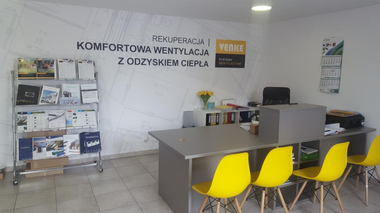 Biuro firmy z logo 'VENKE Systemy Wentylacyjne' na ścianie, ekspozytorem z broszurami i katalogami, szarym biurkiem oraz żółtymi krzesłami w tle ściana z naniesionym planem budynku.