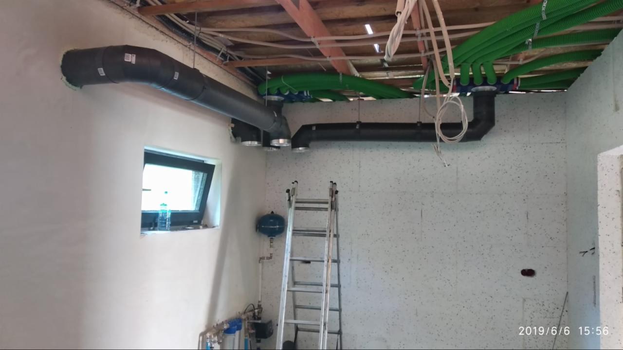 Wnętrze w trakcie montażu systemu rur wentylacyjnych i rekuperacyjnych, widoczne czarne i zielone rury pod sufitem, drabina oparta o ścianę, okno z butelką wody w tle.