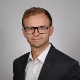 Portret mężczyzny w okularach i czarnej marynarce na szarym tle. Widoczny fragment białej koszuli i czerwony akcent na klapie marynarki. Uśmiechnięty, profesjonalny wygląd.