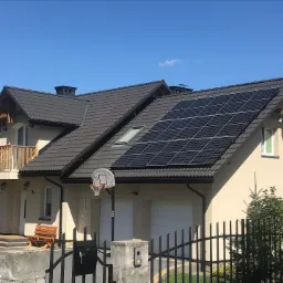 Dom jednorodzinny z panelami słonecznymi na dachu, widok zza czarnego metalowego ogrodzenia z koszem do koszyk&oacute;wki obok garażu.