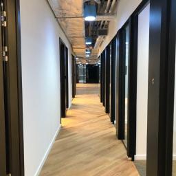 TRADING sp. z o.o. - Długi korytarz biurowy z parkietem ułożonym we wzór jodełki francuskiej, czarne ramy drzwi i odsłonięta instalacja sufitowa z niebieskimi osłonami lamp.