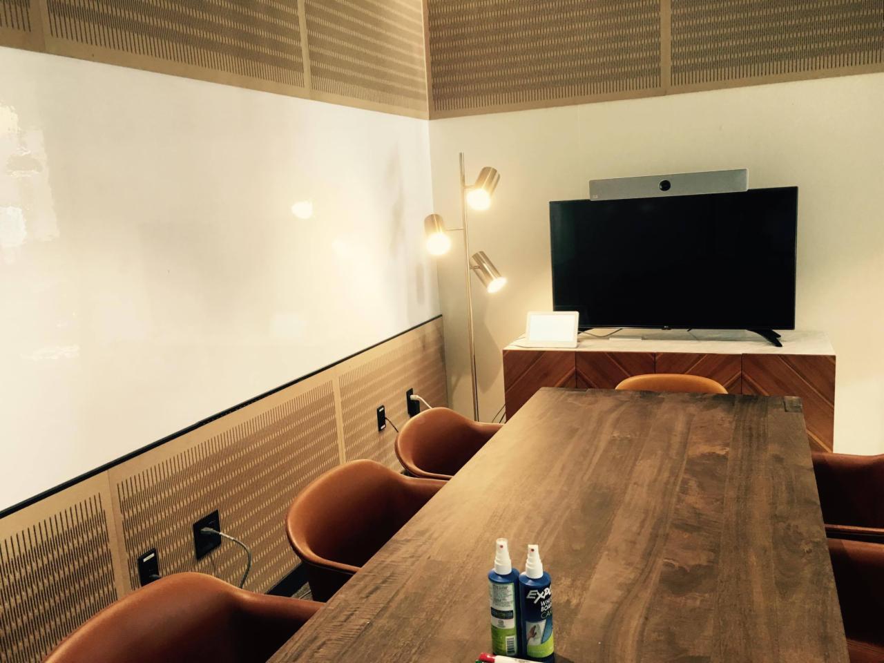 Nowoczesna sala konferencyjna z dużą, drewnianą stołem, białymi ścianami z elementami dekoracyjnymi i telewizorem na szafce. Na stole widoczne środki czystości.