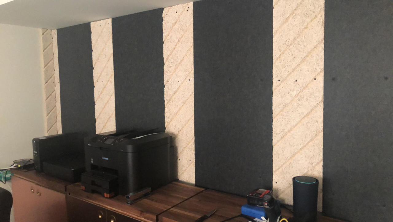 Ciemna ściana z pionowymi pasami z surowego drewna, mocowanymi na wkręty, w tle biurko z drukarkami i głośnikiem.