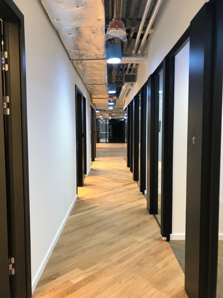 Długi korytarz biurowy z parkietem ułożonym we wzór jodełki francuskiej, czarne ramy drzwi i odsłonięta instalacja sufitowa z niebieskimi osłonami lamp.