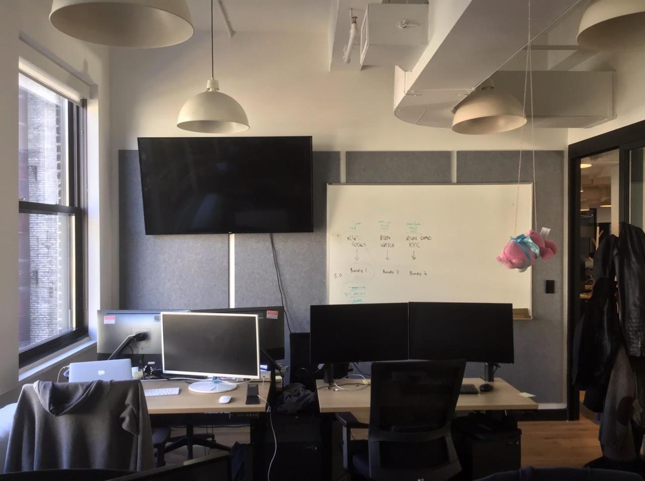 Biuro z dwoma stanowiskami pracy z komputerami, tablicą suchościeralną z zapiskami i wiszącą pluszową świnką, oświetlone lampami wiszącymi.