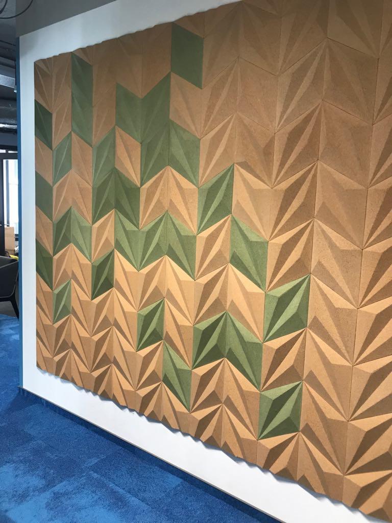 Dekoracyjna ściana z trójwymiarowych paneli w odcieniach brązu i zieleni, ułożonych w geometryczny wzór na białej ścianie, z fragmentem niebieskiej wykładziny w tle.