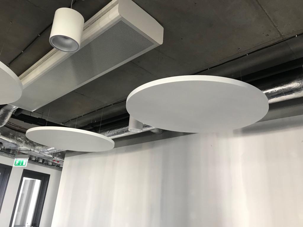 Białe, okrągłe panele akustyczne zawieszone pod betonowym sufitem w biurze, widoczne rury wentylacyjne i oświetlenie