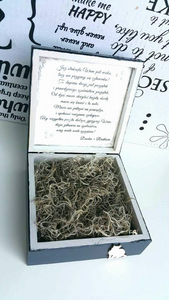 Otwarta, ręcznie robiona, drewniana szkatułka w odcieniach szarości i bieli, wypełniona suszem roślinnym, z wklejoną kartką z życzeniami wewnątrz wieczka, na tle dekoracyjnej tablicy z napisem...