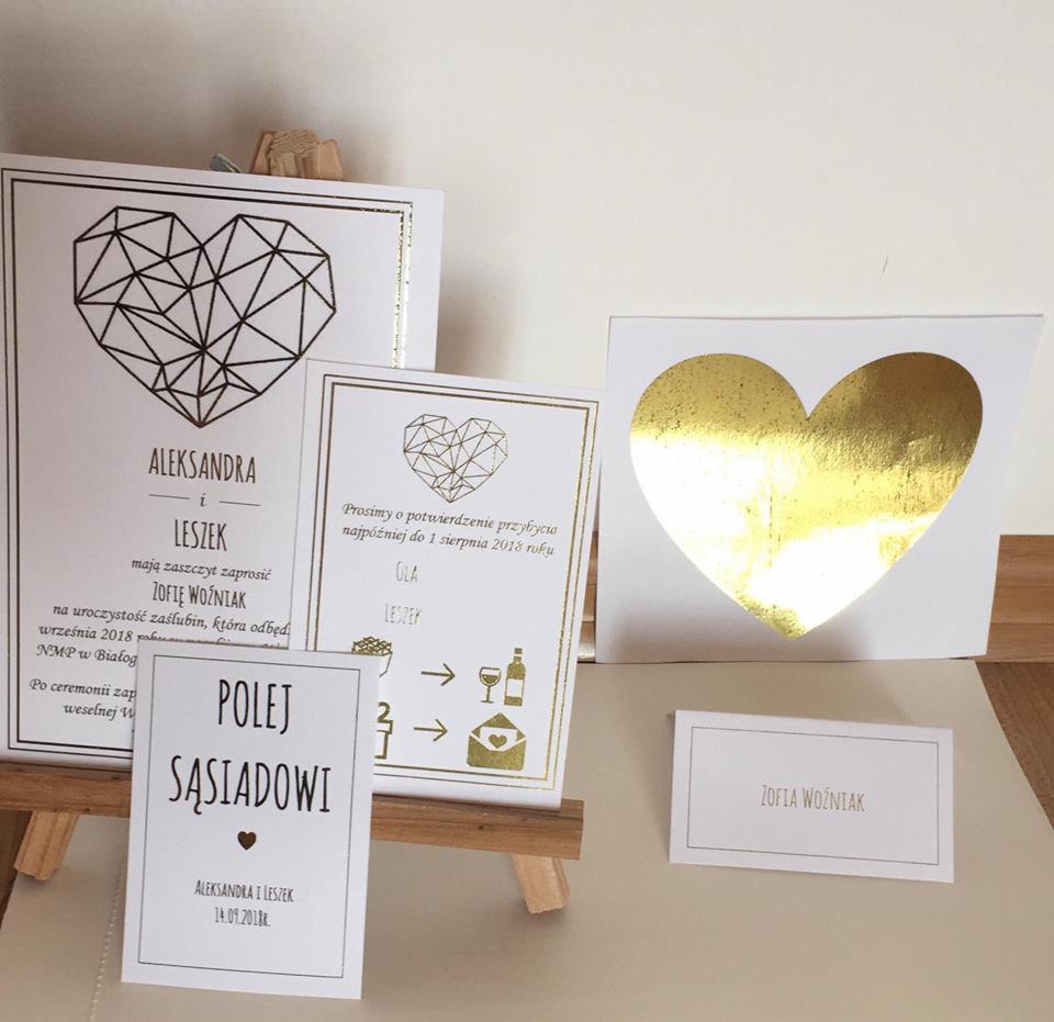 Zestaw złotych i białych zaproszeń ślubnych oraz winietek na drewnianym mini-sztaludze, z motywem serca w różnych wariantach graficznych i typograficznych.