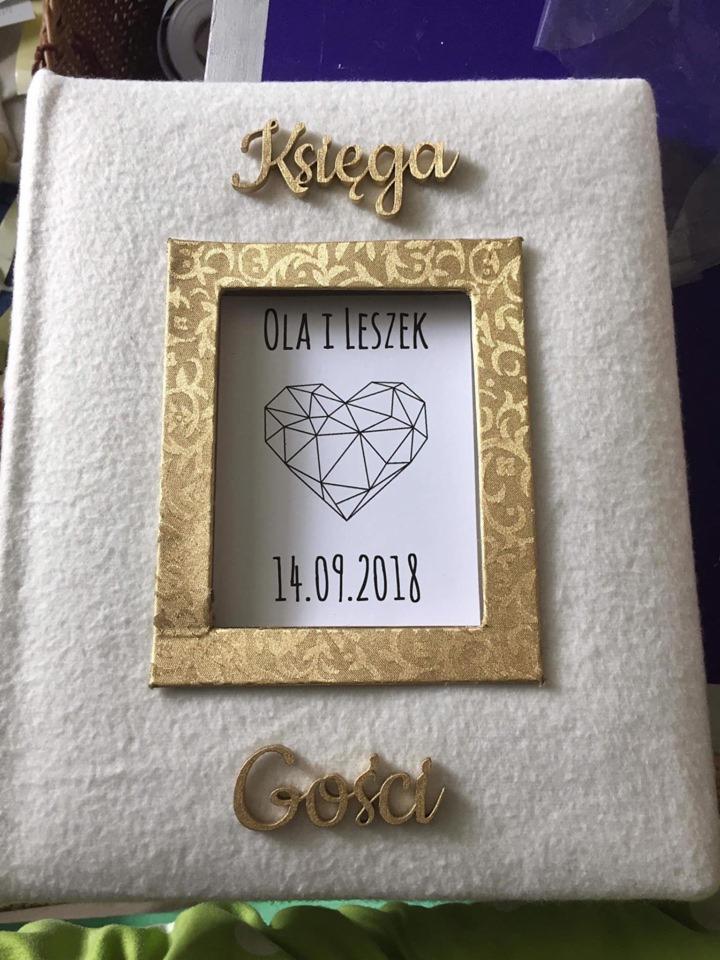 Ręcznie robiona księga gości z białego filcu, ozdobiona złotymi, drewnianymi napisami 'Księga' i 'Gości', ramką ze wzorem oraz kartką z imionami 'Ola i Leszek', datą 14.09.2018 i geometrycznym sercem.