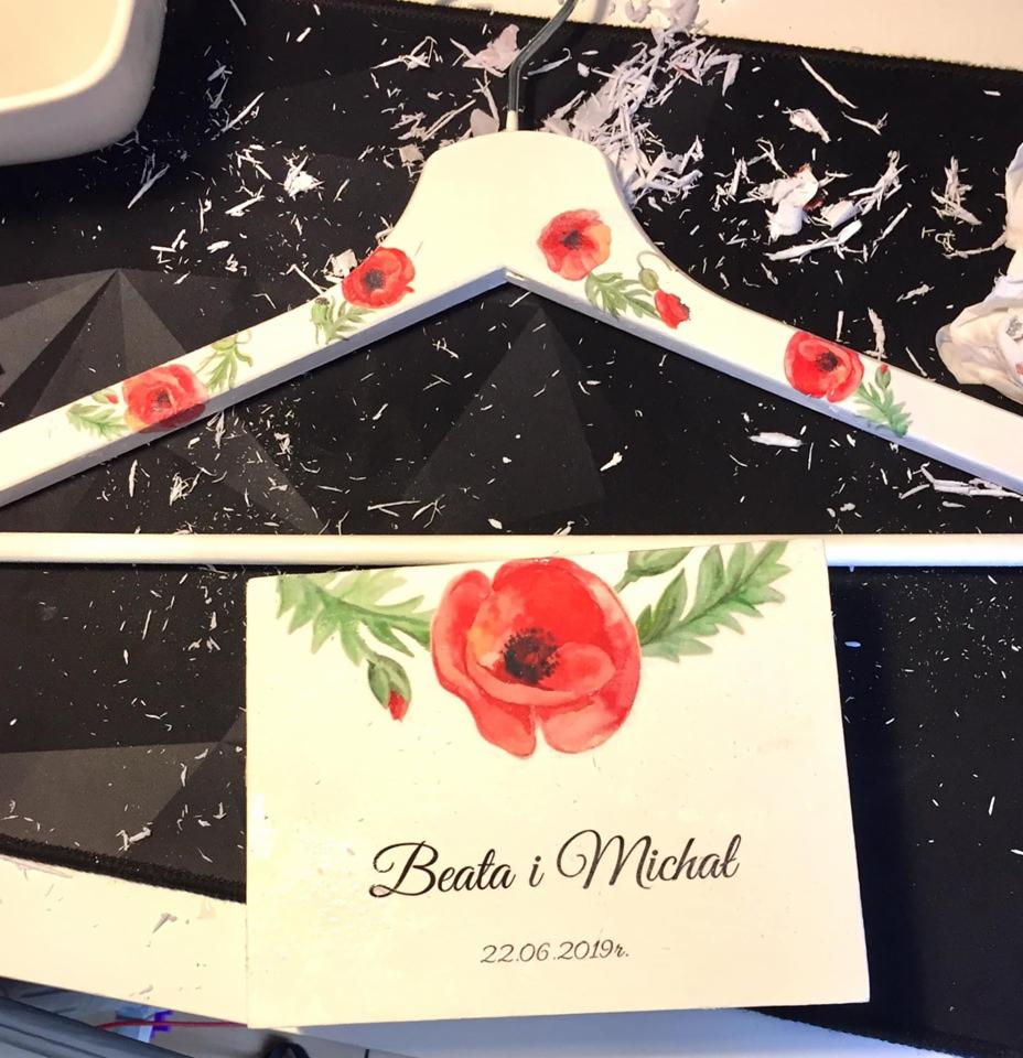 Biały, ręcznie zdobiony wieszak na ubrania z motywem czerwonych maków i kartka z imionami Beata i Michał oraz datą 22.06.2019 na czarnym tle z białymi drobinkami.