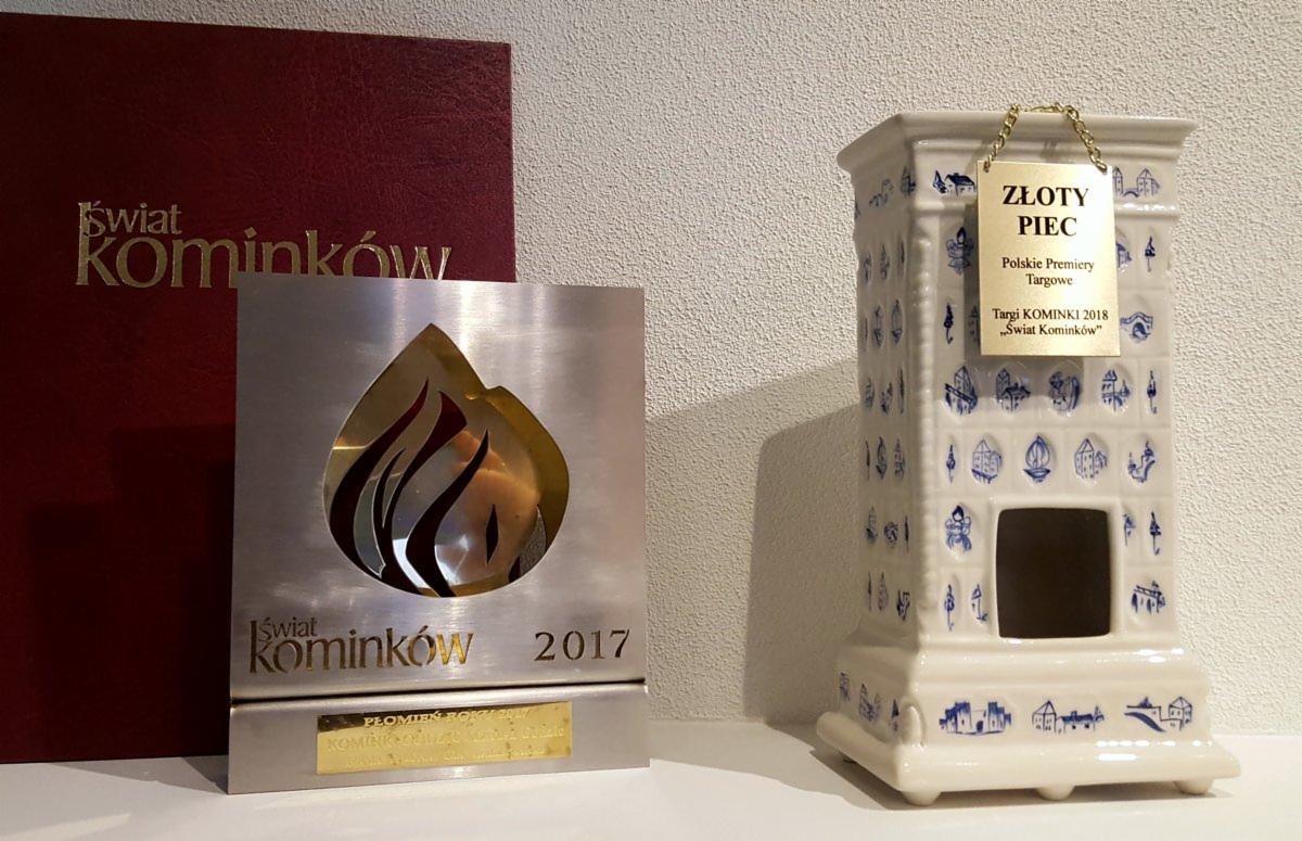 Nagrody i statuetka 'Świat Kominków' z lat 2017 i 2018, obok ceramiczny piec kaflowy z niebieskimi zdobieniami i złotym napisem 'Złoty Piec'.