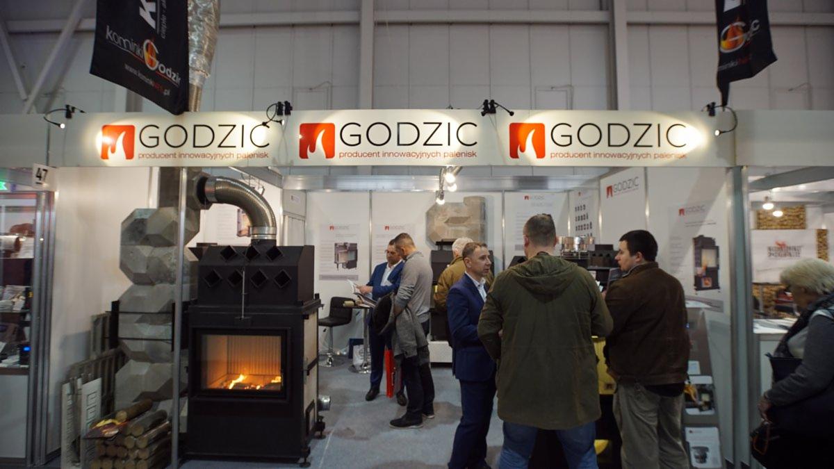 Stoisko targowe producenta palenisk Godzik z czarnym kominkiem z paleniskiem i betonową obudową, widoczne osoby oglądające ekspozycję.
