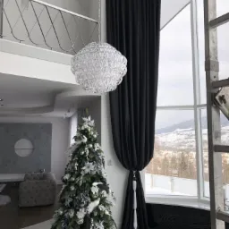 Wysokie okno z czarnymi zasłonami, upięte białym sznurem, obok choinka w odcieniach bieli i srebra, a w tle zimowy krajobraz górski.