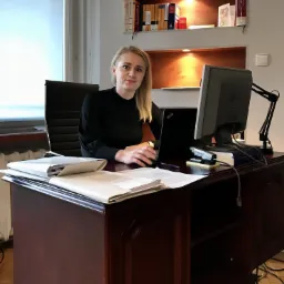 Adwokatka siedzi przy biurku z laptopem i stosem dokumentów, w tle półki z książkami w gabinecie.