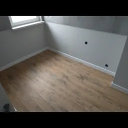 Jasnobrązowe panele podłogowe ułożone w małym, jasnym pokoju z białymi ścianami i szarą górną częścią ściany, widoczne otwory pod gniazdka.