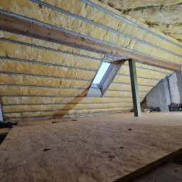 Poddasze w trakcie izolacji, widoczne metalowe profile, wełna mineralna oraz okno dachowe, na podłodze płyta OSB.