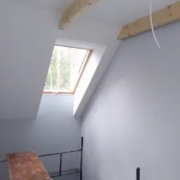 Poddasze po malowaniu na szaro, widoczne drewniane belki stropowe, okno dachowe z widokiem na drzewa oraz fragment rusztowania malarskiego z drewnianą deską.