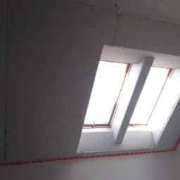 Montaż płyt karton gips na poddaszu wokół okna dachowego, widoczne wkręty i taśma miernicza zabezpieczająca krawędzie.