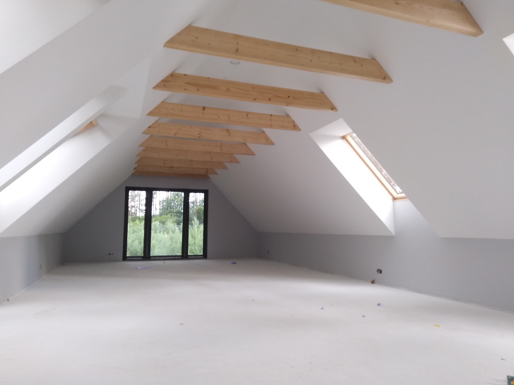 Poddasze po remoncie z drewnianymi belkami podtrzymującymi dach, widoczne okno dachowe i duże okno szklarniowe w szczycie, surowe ściany i podłoga przygotowane do dalszych prac.