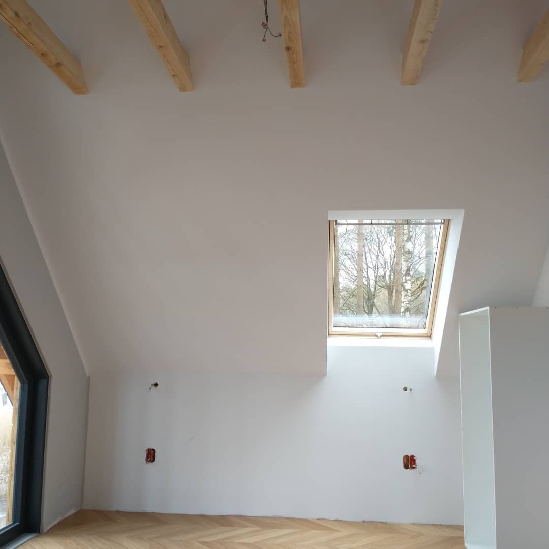 Poddasze po remoncie z oknem dachowym, drewnianymi belkami stropowymi i parkietem w jodełkę; widoczne gniazdka elektryczne i wyprowadzone przewody.