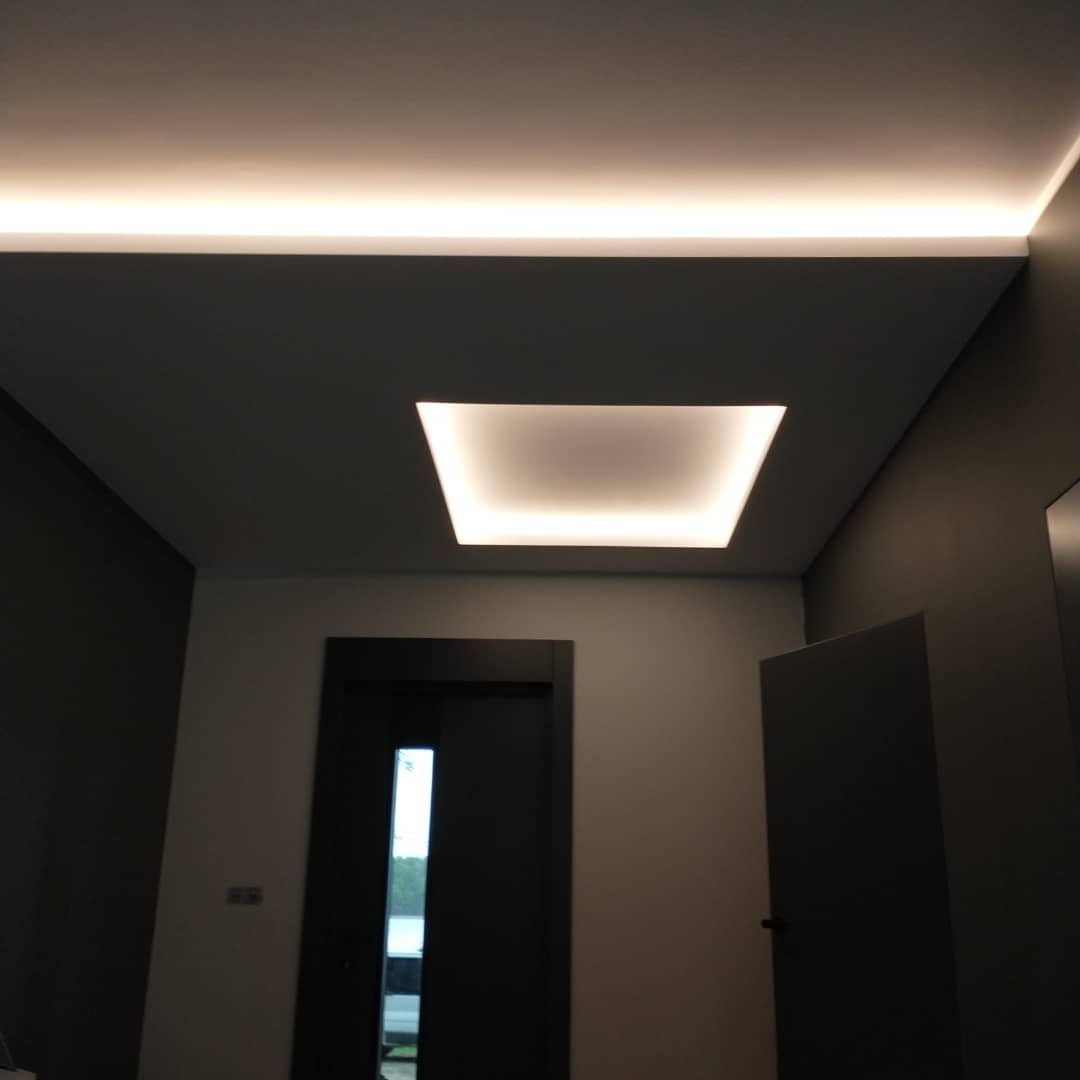 Nowoczesny przedpokój z podwieszanym sufitem, oświetlenie LED w formie prostokątnej wnęki i listwy, ciemne drzwi i ściany w odcieniach szarości.