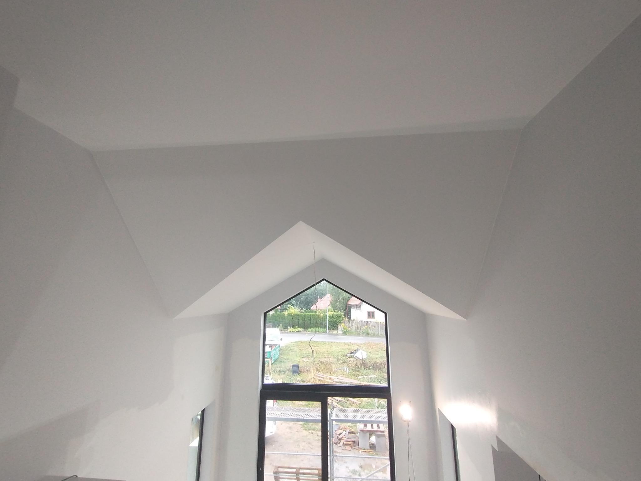 Wnętrze z wysokim sufitem w kształcie litery A, świeżo pomalowane na biało, z dużym oknem w kształcie trójkąta, przez które widać zewnętrzny krajobraz.
