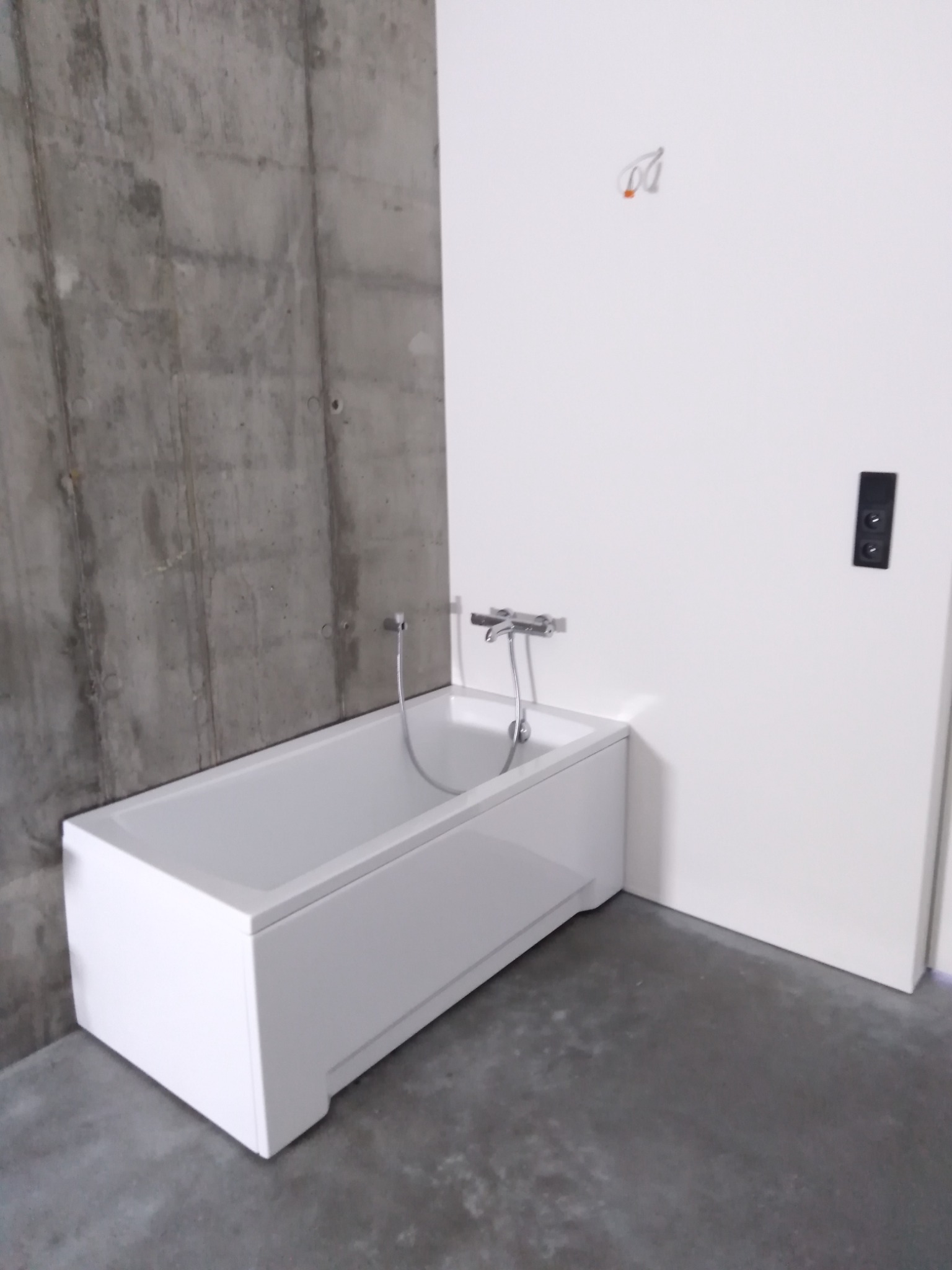Nowoczesna biała wanna z chromowaną baterią, ustawiona przy surowej, betonowej ścianie i gładkiej, białej ścianie z czarnym włącznikiem światła, na szarej, betonowej podłodze.