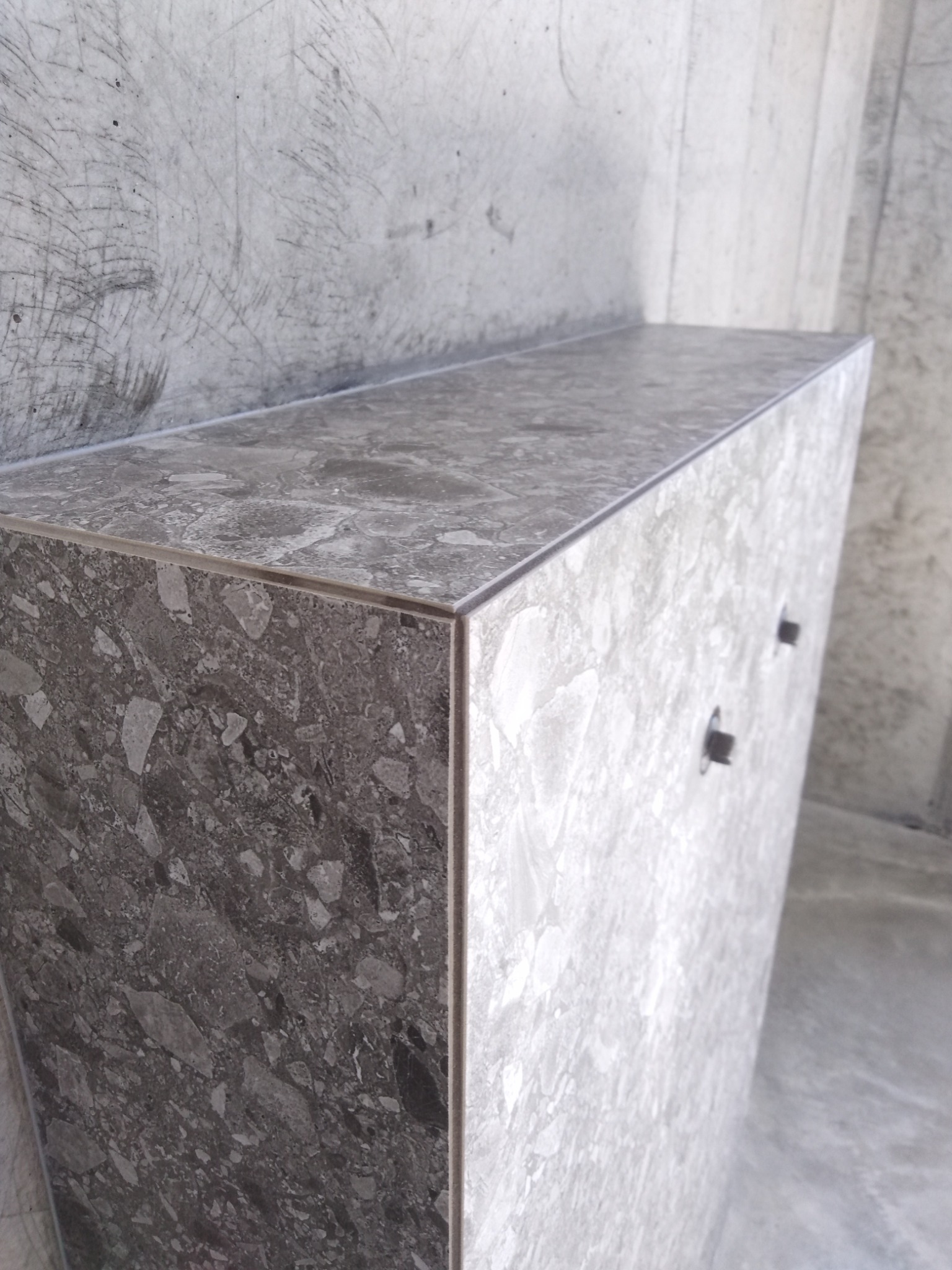 Szara szafka z marmurowym wzorem, minimalistyczny design, dwie metalowe gałki, surowy beton w tle.