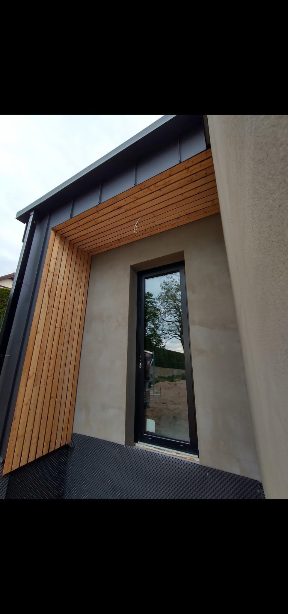 Nowoczesna elewacja domu z drewnianą okładziną, czarną blachą i oknem z czarną ramą; widoczna membrana hydroizolacyjna u podstawy ściany.
