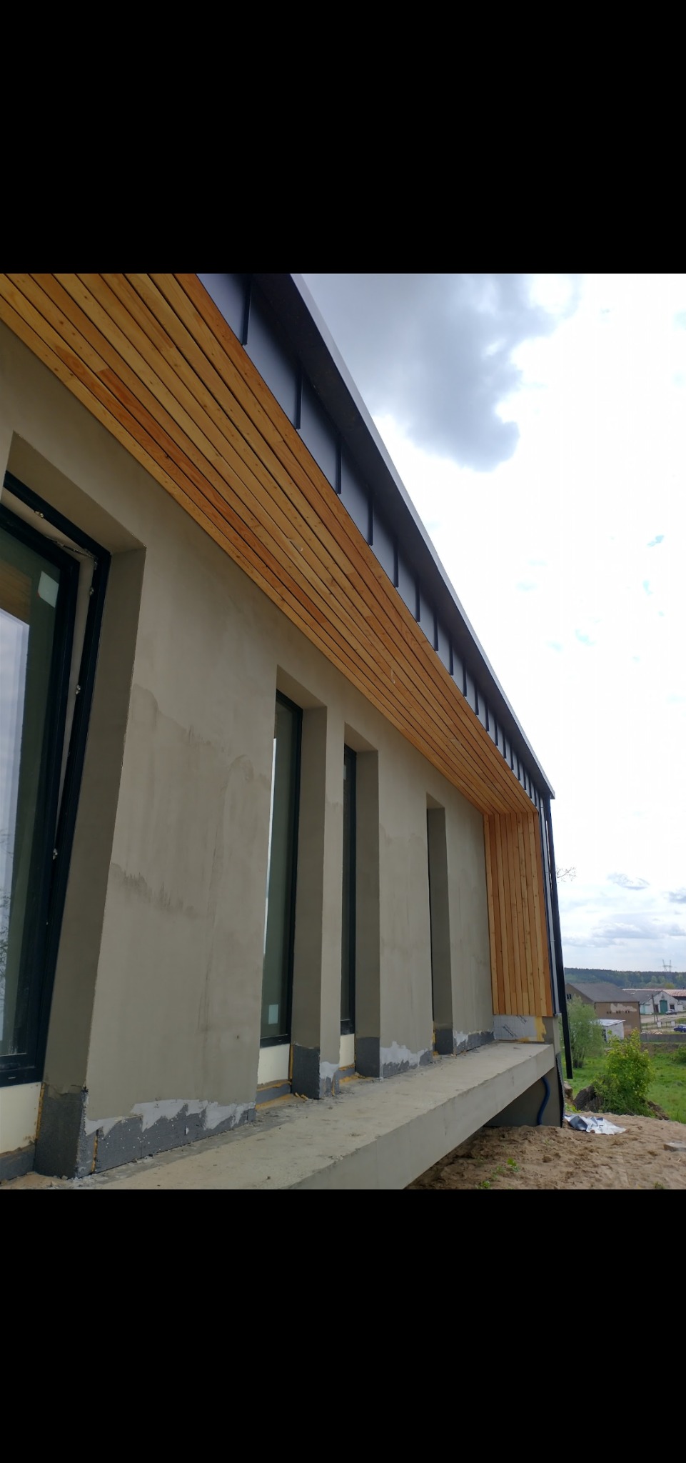 Elewacja budynku w trakcie prac wykończeniowych: surowy tynk, drewniane deski pod okapem i czarne ramy okienne.