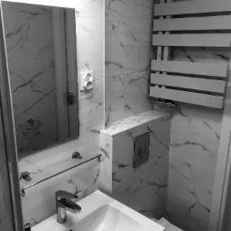 Nowoczesna, monochromatyczna łazienka z marmurowymi płytkami, wiszącą toaletą i grzejnikiem drabinkowym, oświetlona podłużną lampą nad lustrem.