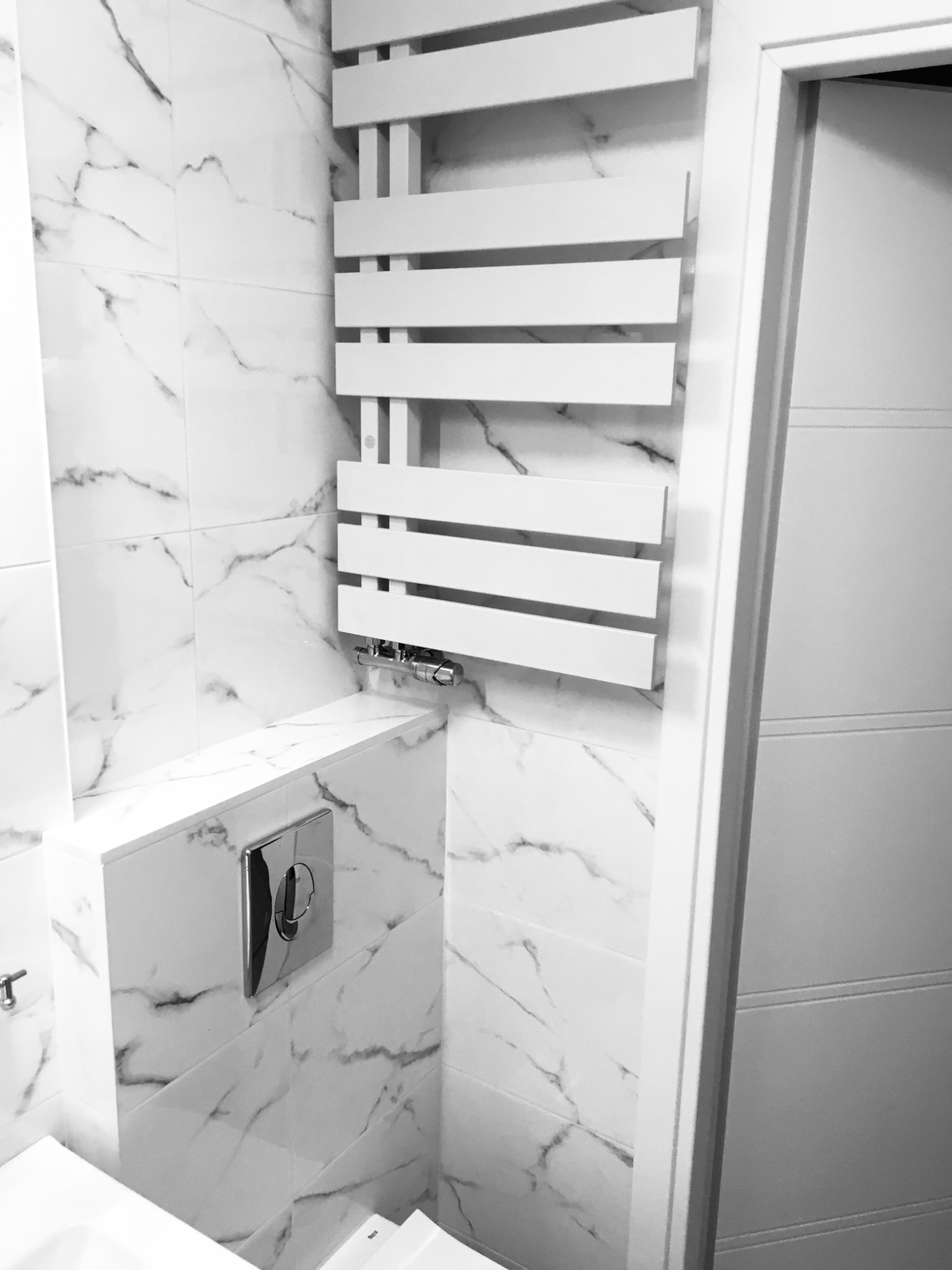 Biała łazienka z marmurowymi płytkami, grzejnikiem drabinkowym i podtynkową spłuczką WC w chromowanej obudowie.