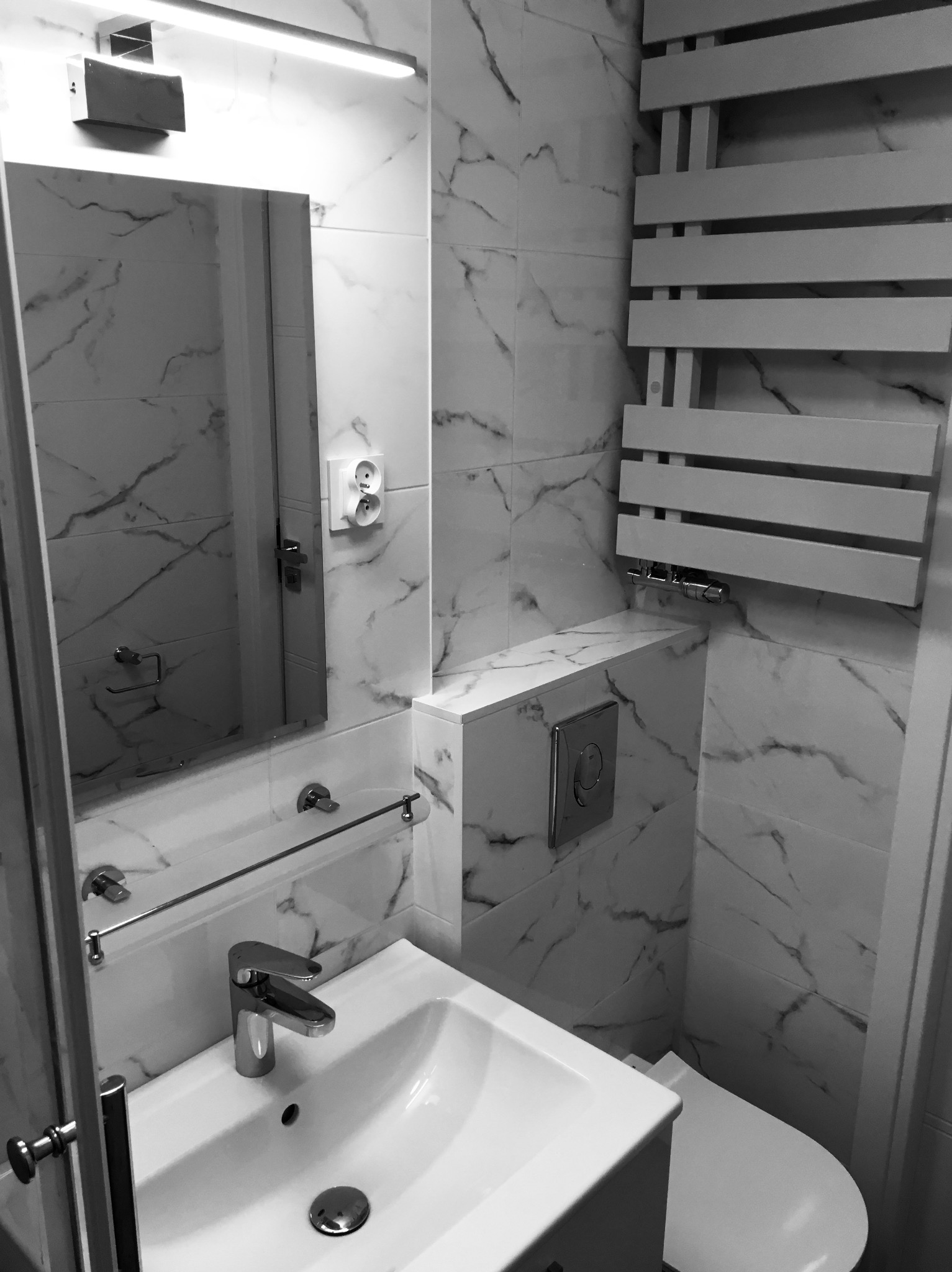 Nowoczesna, monochromatyczna łazienka z marmurowymi płytkami, wiszącą toaletą i grzejnikiem drabinkowym, oświetlona podłużną lampą nad lustrem.