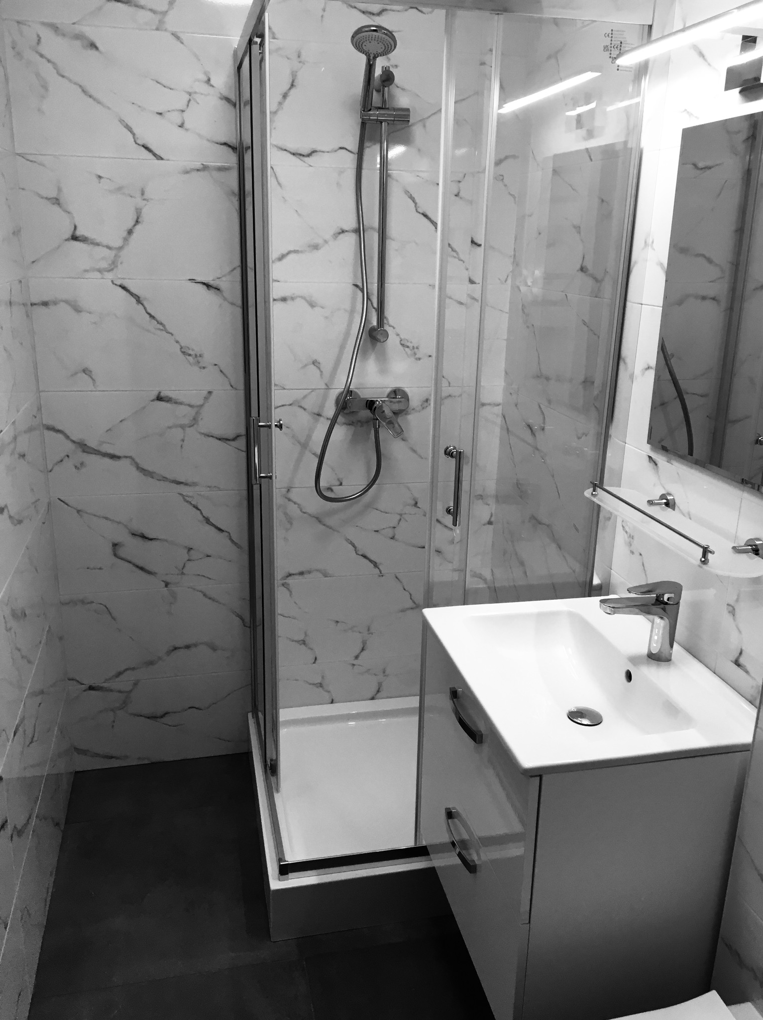 Wykończona łazienka w czerni i bieli: kabina prysznicowa z przesuwnymi drzwiami, umywalka z szafką i marmurowe płytki ścienne z wyraźnym wzorem żyłkowania.