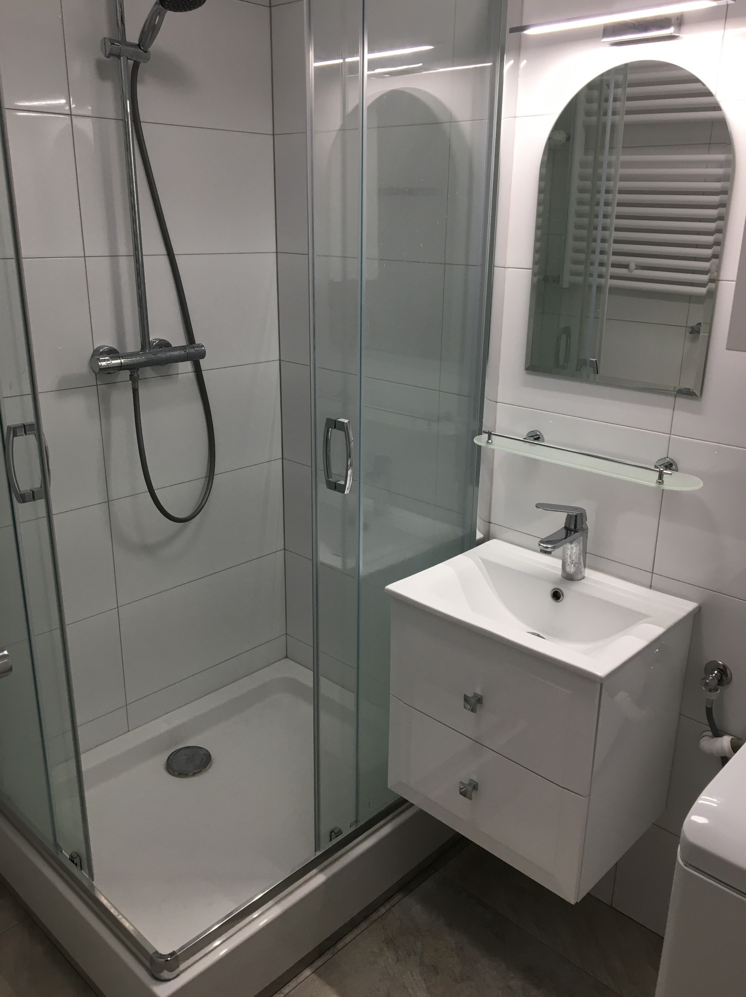 Nowoczesna, biała łazienka z kabiną prysznicową ze szklanymi drzwiami, umywalką z szafką i owalnym lustrem podświetlonym od góry, na ścianach białe płytki.