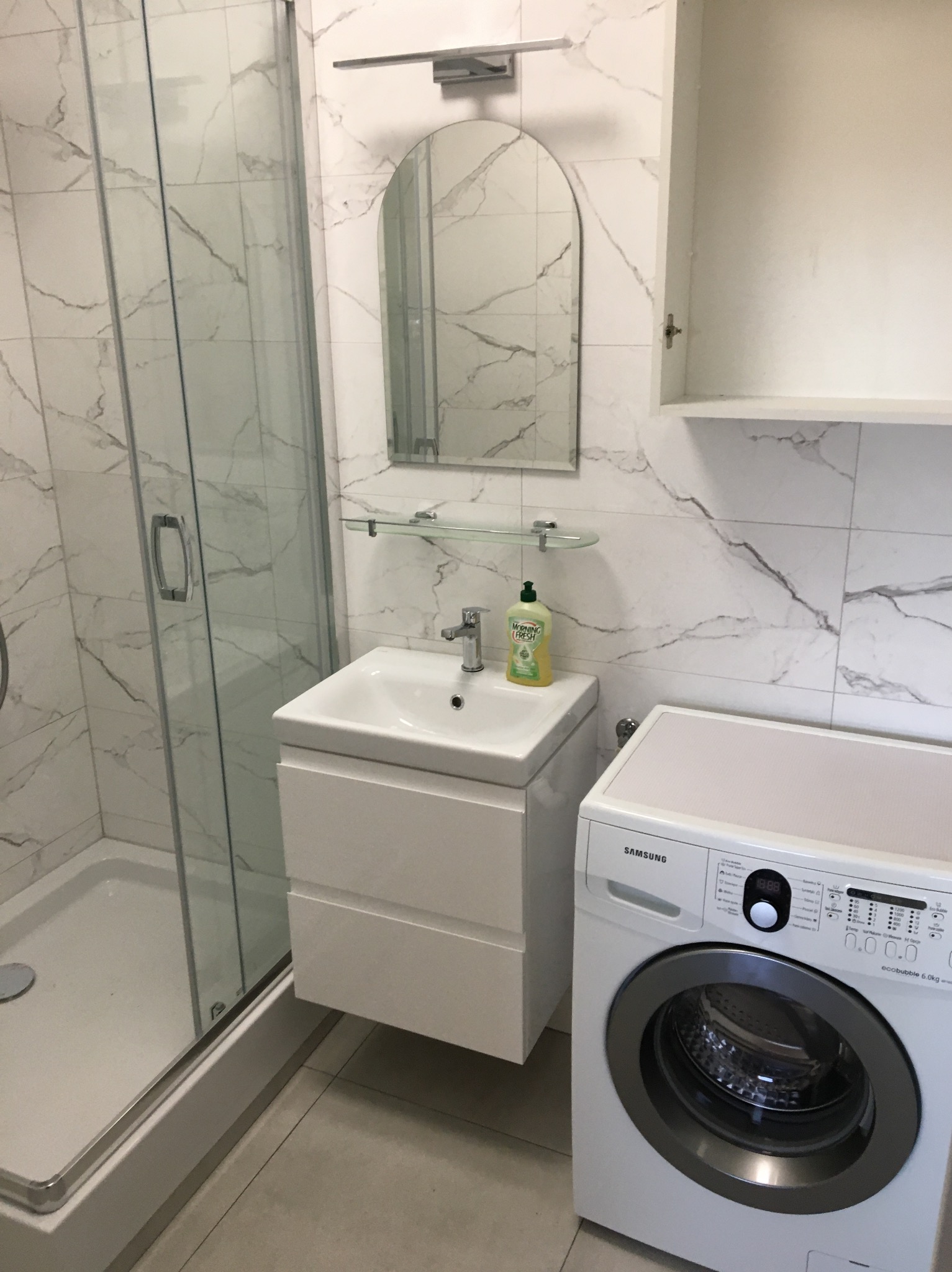 Biała łazienka z kabiną prysznicową, umywalką z szafką, owalnym lustrem, szafką wiszącą i pralką Samsung Ecobubble 6.0 kg.