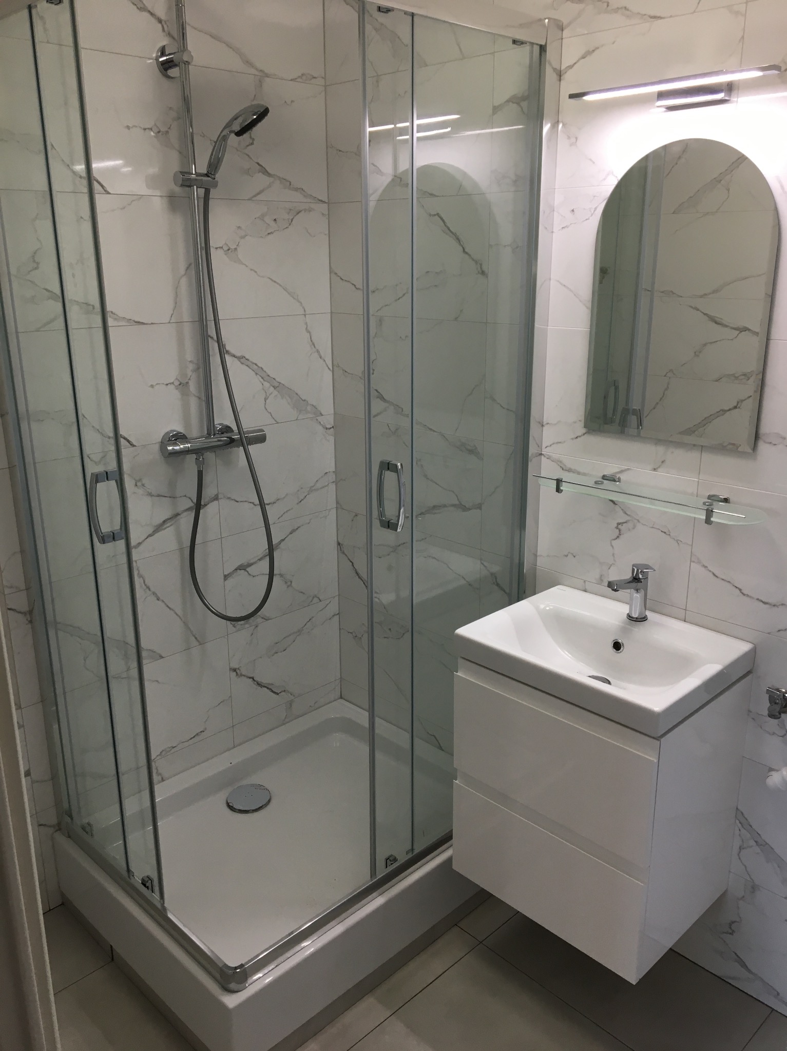 Nowoczesna łazienka z kabiną prysznicową z przezroczystego szkła, umywalką na szafce z dwiema szufladami i lustrem z oświetleniem LED na ścianie wyłożonej płytkami imitującymi marmur.