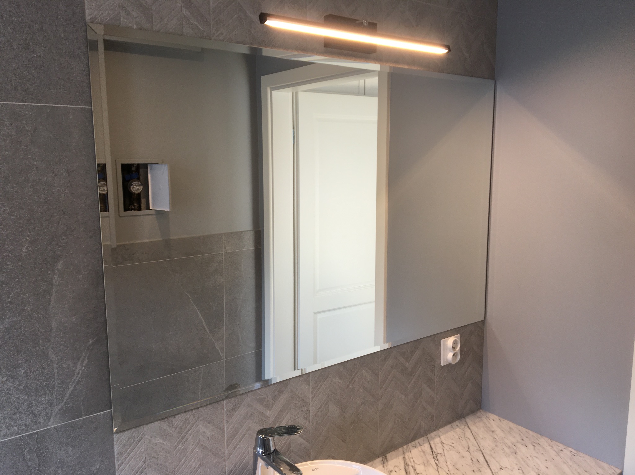 Nowoczesna łazienka z dużym lustrem podświetlonym lampą LED, umywalką z marmurowym blatem i szarą płytką imitującą kamień, widoczny fragment drzwi w odbiciu lustra.