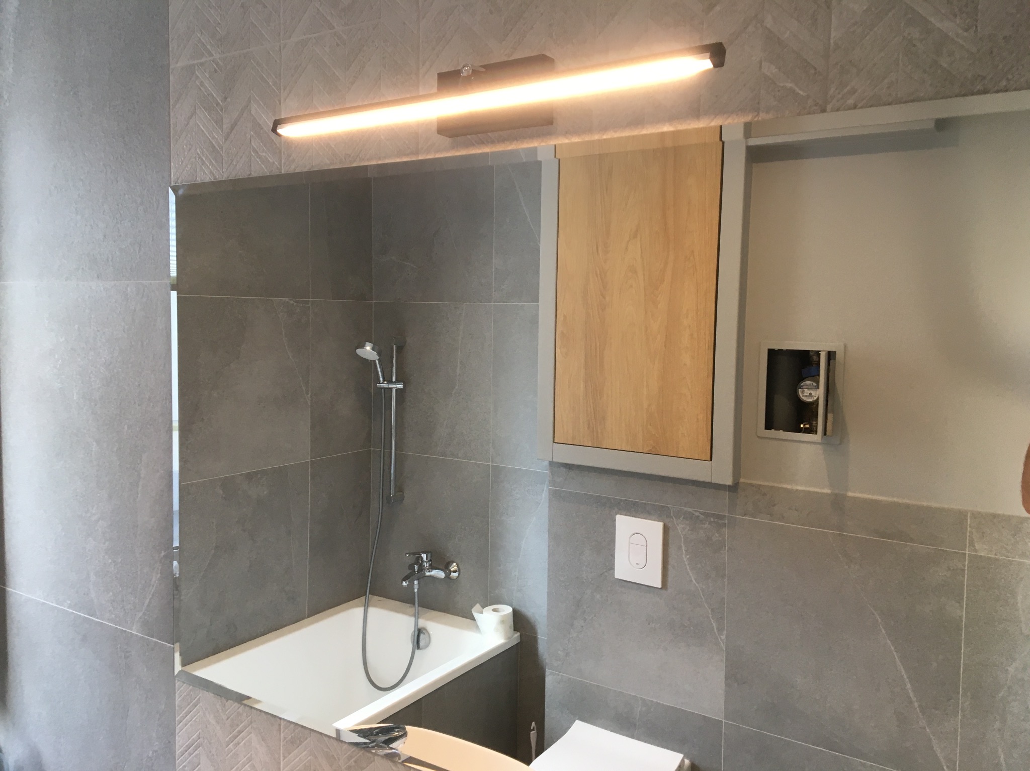 Nowoczesna łazienka z szarymi płytkami w jodełkę, podświetlanym lustrem i drewnianą szafką w zabudowie, widoczna toaleta i wanna z baterią.