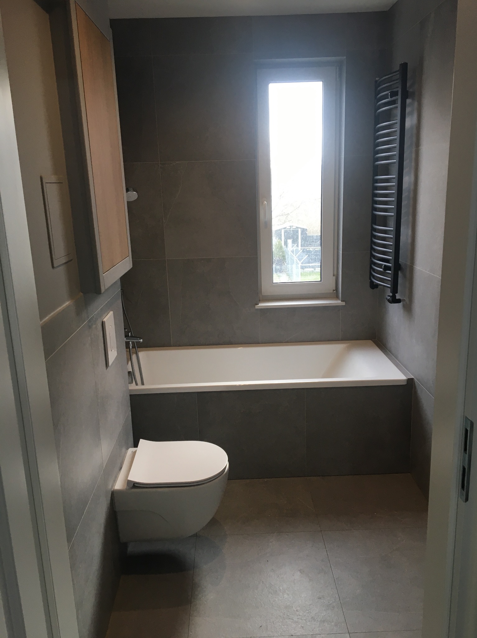 Nowoczesna łazienka z szarymi płytkami, podwieszaną toaletą, białą wanną pod oknem i grzejnikiem drabinkowym w kolorze czarnym.