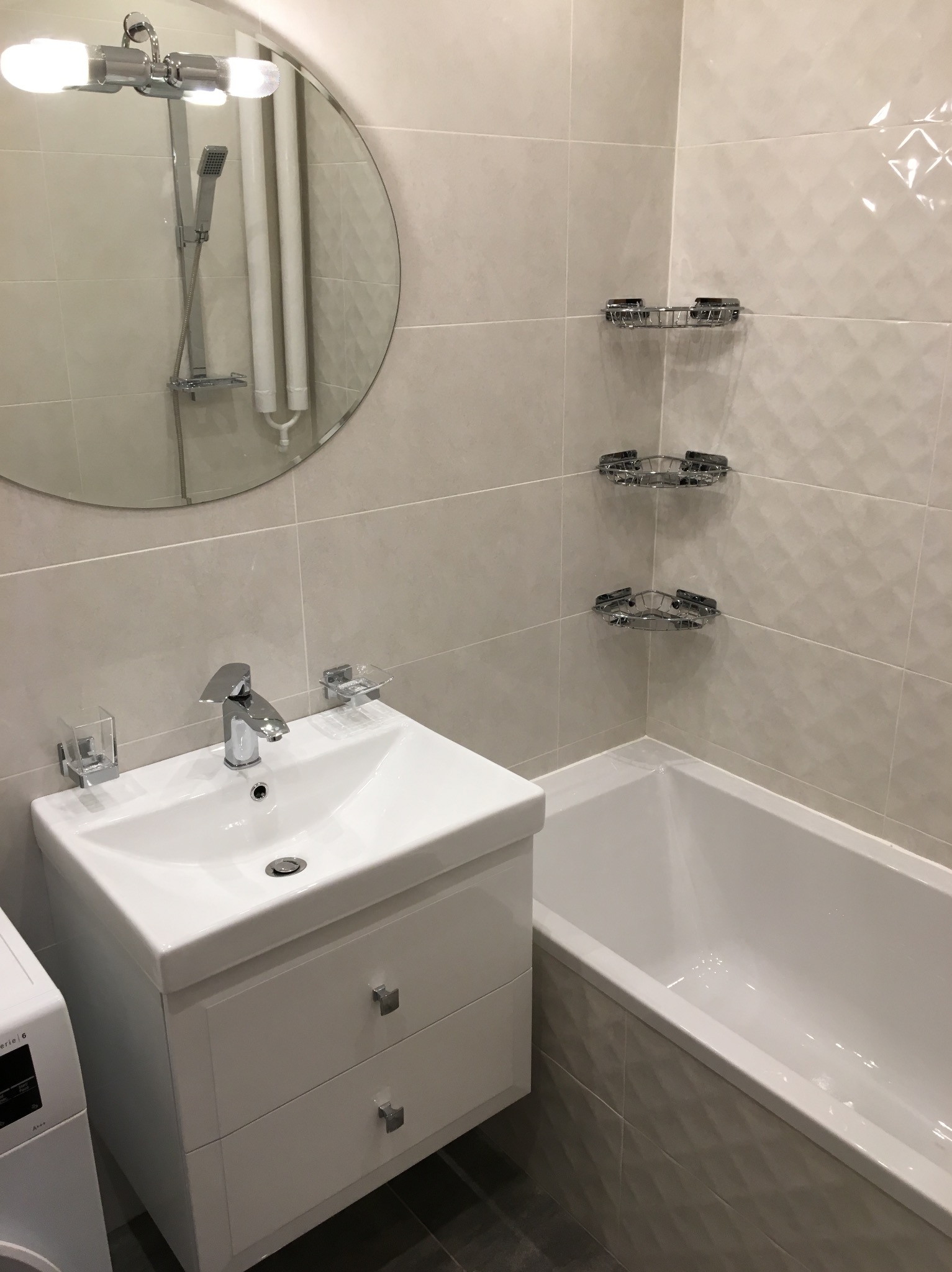 Wykończona łazienka w jasnych odcieniach z owalnym lustrem, białą umywalką z szafką, wanną obudowaną płytkami z fakturą oraz chromowanymi półkami narożnymi.