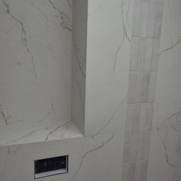Dawid Gol Usługi Remontowo-Budowlane - Wykończona ściana w łazience z płytkami imitującymi marmur z delikatnym wzorem żyłkowania i wstawką z płytek w innym odcieniu. Widoczny otwór na gniazdko.